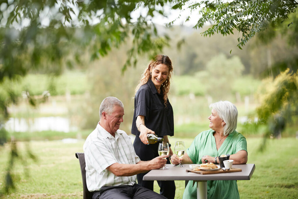 older couple sitting outdoors under and near trees by lake with waitress pouring drink