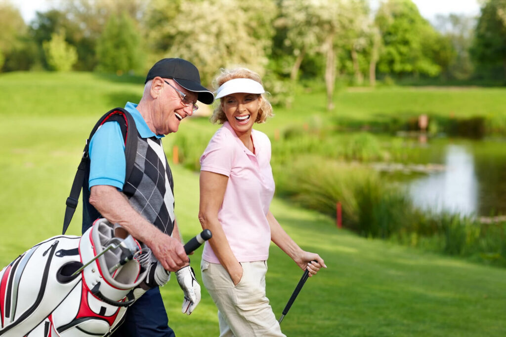 older lady and man walking on golf green with man carrying clubs and woman holding a golf stick mid stride