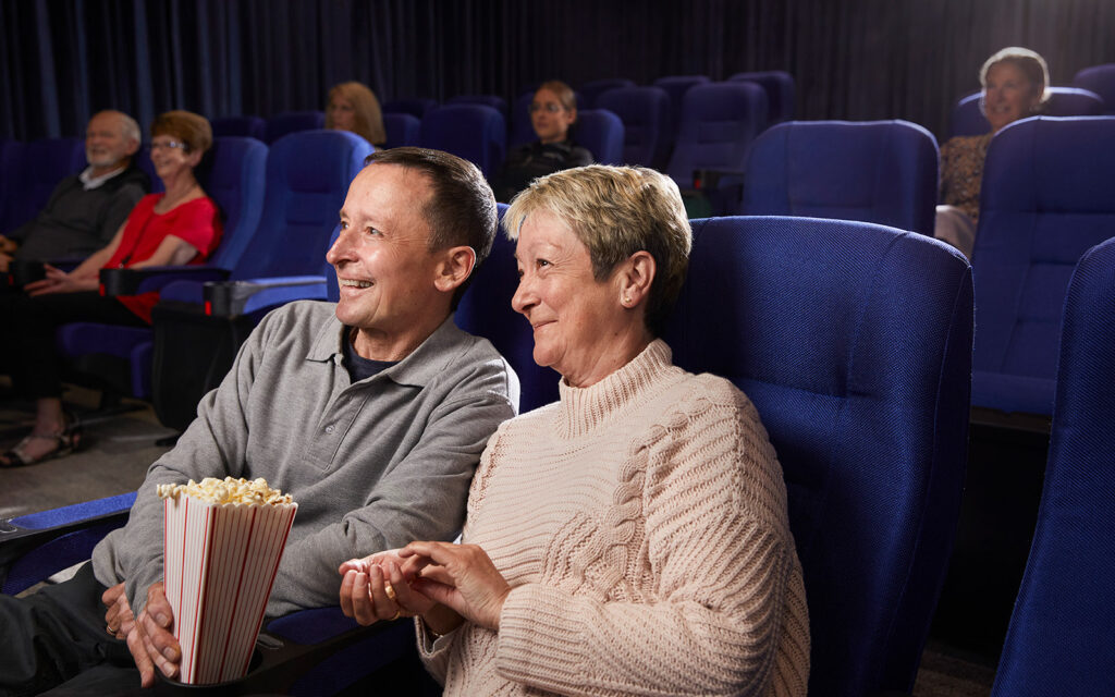 older lady and gentleman sitting in cinema chairs with other people randomly seated in the background